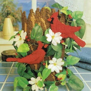 May include: A brown and tan woven vase with two red cardinal birds and white flowers. The vase is surrounded by green leaves.