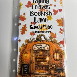 May include: A clear plastic binder insert featuring a white background with orange and yellow autumn leaves. The text reads Falling Leaves Bookish Lane Saves $550 BudgetingWithKim. Below is a large orange pumpkin shaped bookstore with a sign reading Pumpkin Reads Bookstore and a small sign reading Books Are Magic.