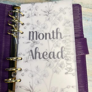 May include: A purple planner with gold rings and a clear overlay. The overlay features a floral design and the words "Month Ahead" in a script font. The planner is made of a textured material.