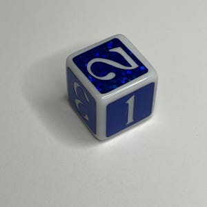 May include: A six-sided die with a white frame and blue faces. The top face features a glittery blue surface with the white number 2, while the front face displays the white number 1 on a solid blue background. The side face shows a partial white number 3.