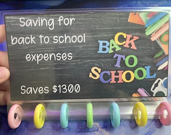 May include: A chalkboard sign with the text "Saving for back to school expenses" and "Saves $1300". The sign also has the words "Back to School" in colorful letters. There are six colorful rings hanging below the sign.