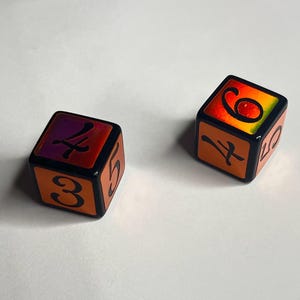 May include: Two six-sided dice with black edges and orange faces. The top faces feature a gradient of red, orange, and yellow with black numbers 4 and 6. The side faces are orange with black numbers 3, 5, 4, and 10 printed in a serif font.
