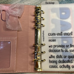 May include: A light pink six-ring binder with a textured surface, a sheer pink ribbon bow, and a gold-tone metal ring mechanism. A translucent divider is inserted, featuring the printed text: Birthdays, BudgetingWithKim, and a definition of commitment.