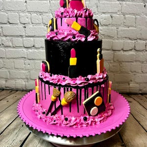 May include: A three-tiered cake decorated with makeup-themed fondant pieces including lipsticks, makeup brushes, and a compact palette. The cake features alternating layers of bright pink and black frosting with black drip accents and pink piping.