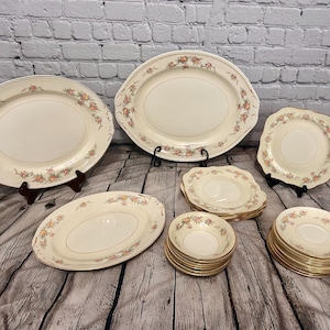 Könnte beinhalten: Eine Sammlung von cremefarbenem Vintage-Geschirr mit einem Blumenmuster in Pfirsich- und Grüntönen. Das Set umfasst ovale Platten, runde Teller und Schalen mit Goldrand, die auf einer Holzoberfläche präsentiert werden.