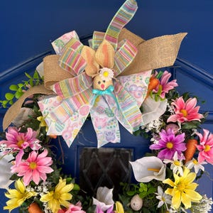 May include: A colorful Easter wreath with pink, yellow, and white flowers, green leaves, and a burlap bow with a bunny figurine in the center.