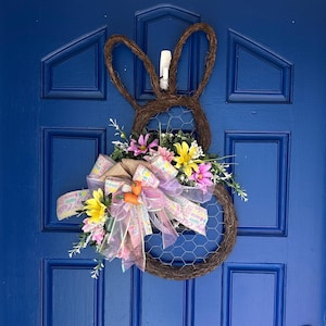 May include: A brown twig bunny wreath with a pink and yellow floral arrangement and a burlap bow with the text "Happy Easter" on it. The wreath is hanging on a blue door.