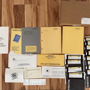 May include: A collection of vintage computer software and documentation, including manuals, floppy disks, and a cardboard box. The items are arranged on a wooden surface, with text visible on the manuals and disk labels.