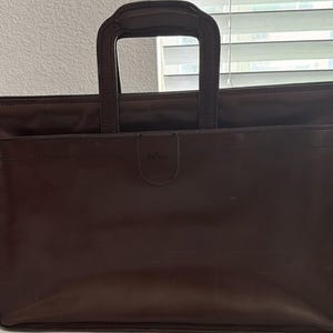 May include: A dark brown leather briefcase featuring a rectangular silhouette, two top handles, and a front exterior pocket with a central flap closure. The brand name 'halson' is embossed on the small tab of the front pocket closure.