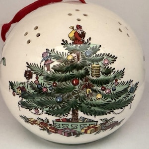 May include: A spherical white ceramic ornament featuring a detailed illustration of a decorated Christmas tree with a figure of Santa Claus at the top, surrounded by colorful ornaments and wrapped gifts at the base, topped with a red ribbon loop.