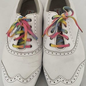 May include: Pair of white leather golf shoes with rainbow-colored laces. The shoes have a classic wingtip design with decorative perforations along the edges. The laces are a vibrant mix of colors.