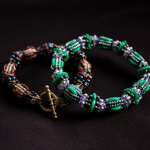 May include: Two beaded bracelets, one with green and silver beads and the other with red, gold, and brown beads. The bracelets are intertwined and have a gold clasp.