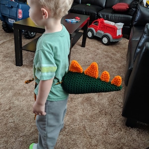 May include: A young child wearing a green shirt and grey trousers with a crocheted dinosaur tail attached to their waist. The tail is green with orange spikes.