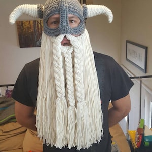 May include: A person wearing a gray knitted Viking helmet with horns and a long white yarn beard.