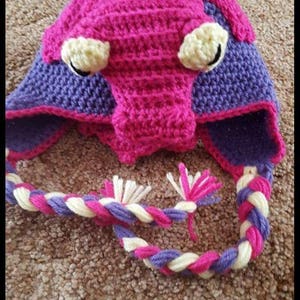 May include: A pink and purple crocheted hat with a long braided tail. The hat has two yellow eyes and a purple nose.