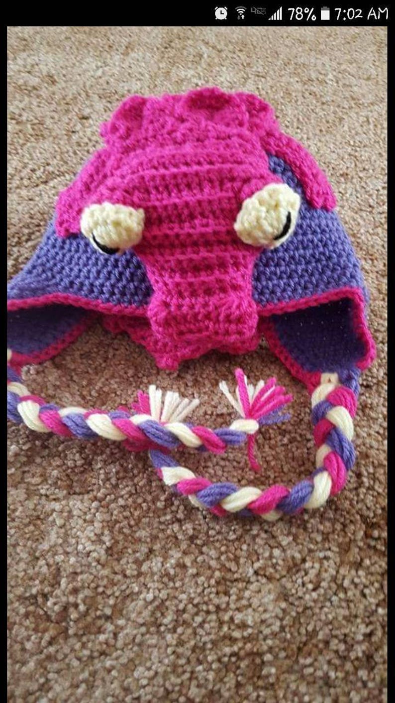 May include: A pink and purple crocheted hat with a long braided tail. The hat has two yellow eyes and a purple nose.