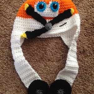 May include: A handmade crochet hat in the shape of an airplane. The hat features an orange top, white body, and black propeller. It has blue eyes and black wheels. The hat is designed to be worn on the head, with ear flaps.