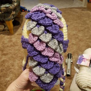 May include: A crocheted hat with a dragon scale design in shades of purple, pink, and gray. The hat has a long tail that is braided with the same colors.