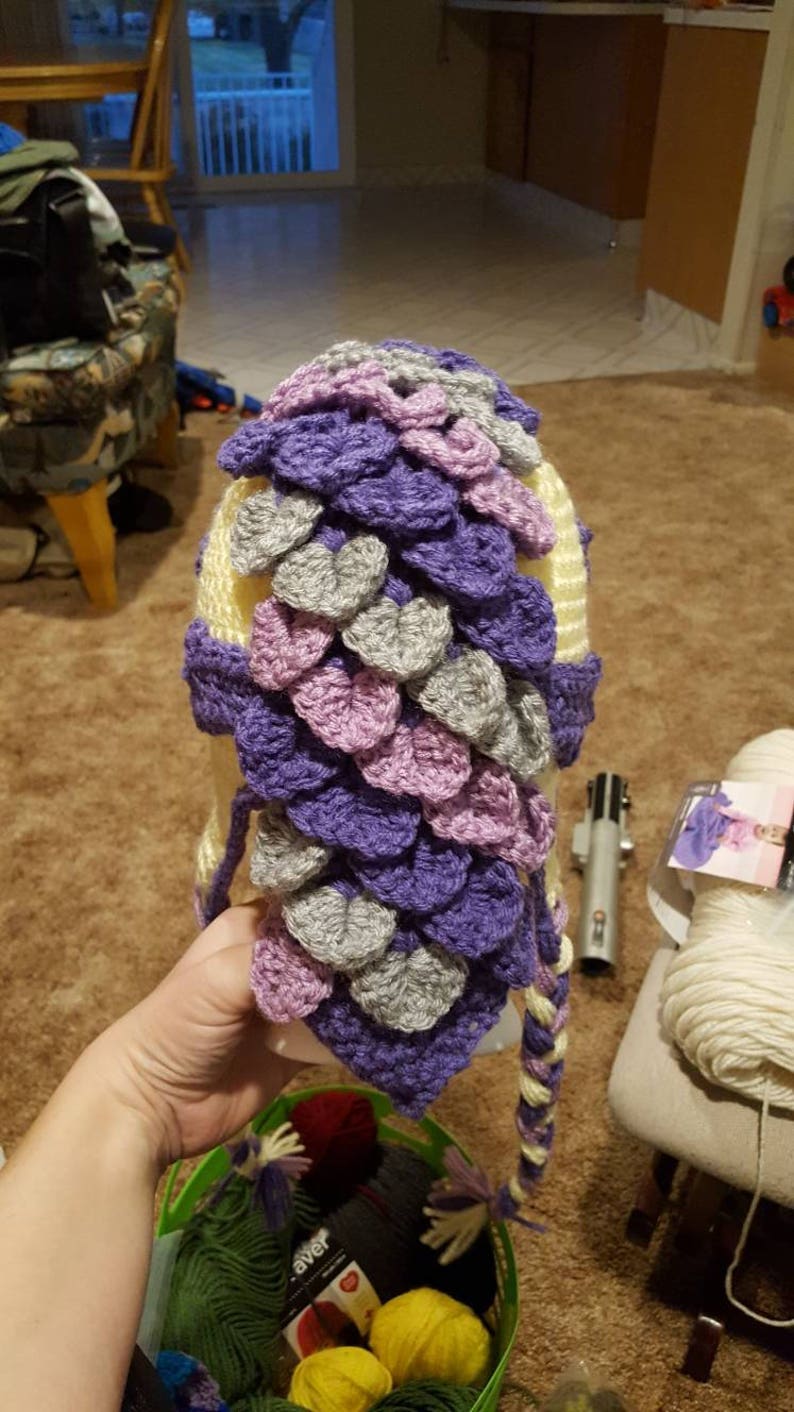 May include: A crocheted hat with a dragon scale design in shades of purple, pink, and gray. The hat has a long tail that is braided with the same colors.