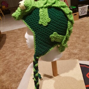 May include: Green crocheted hat shaped like an alligator with a long green and black braided tail. The hat is on a white mannequin head.