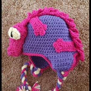 May include: Purple and pink crocheted hat with a long braided strap. The hat is shaped like a cartoon animal with a large eye and a pink snout.