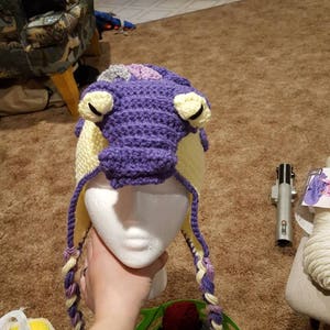 May include: A purple and yellow crocheted hat with a dragon design. The hat is on a white styrofoam head and has long braids.