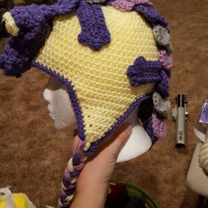 May include: A yellow, purple, and gray crocheted hat with a long braid. The hat is on a white mannequin head.