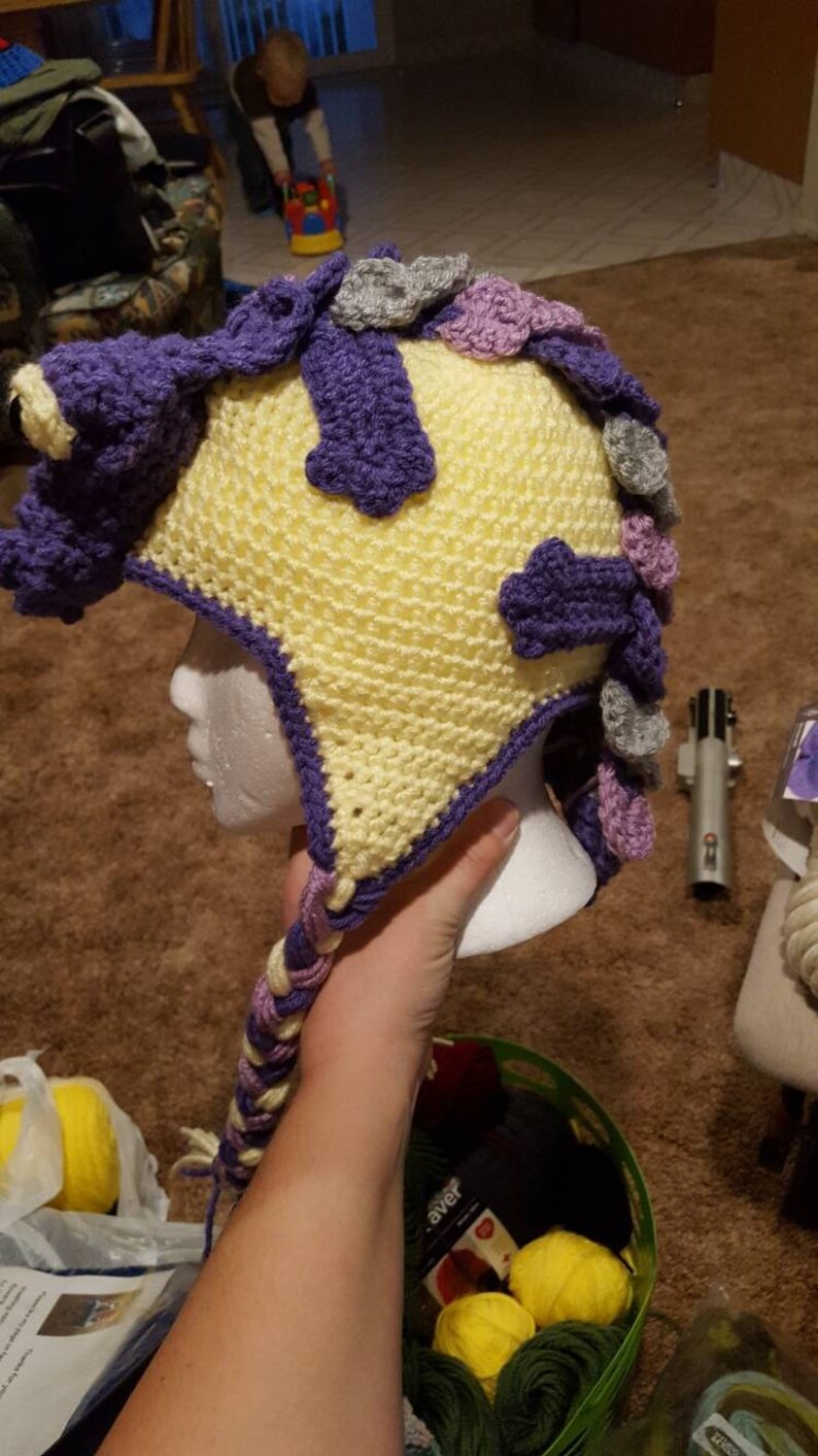May include: A yellow, purple, and gray crocheted hat with a long braid. The hat is on a white mannequin head.