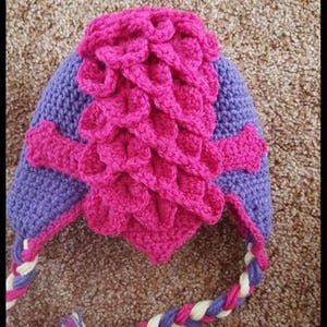 May include: A pink and purple crocheted hat with a braided strap. The hat has a unique design with a textured pattern.