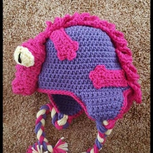 May include: A handmade crochet hat in purple and pink, featuring a whimsical monster design. The hat has a large eye, decorative patches, and a braided tie with tassels. The hat is designed for children.
