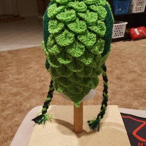 May include: Green and black crocheted hat with a scale pattern and long braids. The hat is displayed on a wooden stand.