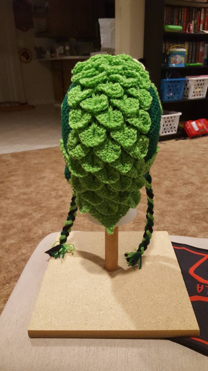 May include: Green and black crocheted hat with a scale pattern and long braids. The hat is displayed on a wooden stand.