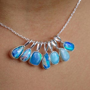 Op de afbeelding: Een zilveren ketting met zeven individuele hangers, elk bezet met een unieke gepolijste opaal edelsteen. De opalen vertonen verschillende tinten blauw, groen en iriserende flitsen, gevat in eenvoudige zilveren zettingen aan de ketting.