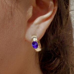 May include: A close-up of a gold-colored hoop earring with a vibrant blue oval-shaped gemstone. The earring is worn on an ear, with brown hair visible in the background. The earring's design is simple and elegant.