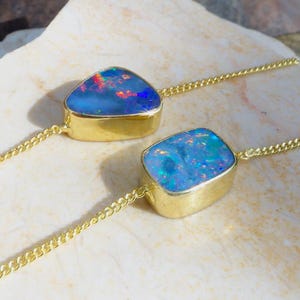 Puede incluir: Dos pulseras de cadena dorada con piedras de ópalo engastadas. Un ópalo es triangular con destellos azules y rojos, mientras que el otro es rectangular con motas iridiscentes azules y verdes. Las cadenas doradas son finas y se unen a los engastes.
