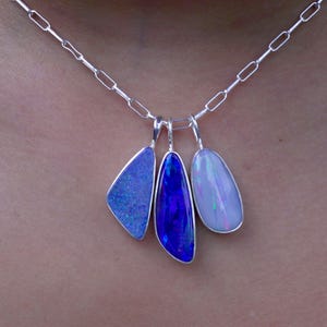 Op de afbeelding: Een zilveren ketting met drie opalen hangers in blauwe en paarse tinten. De hangers hebben een druppelvorm en zijn gezet in zilver. De ketting wordt om de nek gedragen.