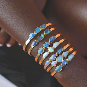 Op de afbeelding: Een stapel van zeven goudkleurige metalen armbanden, elk voorzien van twee onregelmatige, gepolijste opaalstenen. De opalen vertonen iriserende flitsen van blauw, groen en wit, gezet in gouden randen langs de dunne, gladde, metalen banden.
