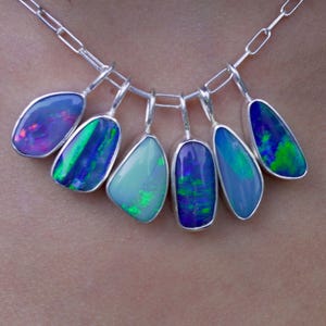 Könnte beinhalten: Eine silberne Halskette mit sieben Opalanhängern. Jeder Anhänger hat eine einzigartige Form und zeigt lebendige Farben, darunter Blau, Grün, Lila und Rosa. Die Opale sind in Silberfassungen gefasst und mit einer Silberkette verbunden.