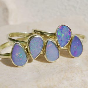 Könnte beinhalten: Zwei Goldringe mit tropfenförmigen Opalsteinen. Jeder Ring verfügt über eine glatte, polierte Goldschiene und einen in einer Zargenfassung gefassten Opal mit hellblauer Grundfarbe und irisierenden rosa, grünen und violetten Lichtreflexen.