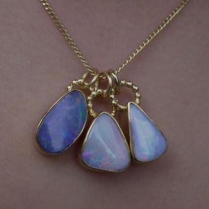 Peut inclure: Un collier à chaîne dorée avec trois pendentifs en opale en forme de goutte. Chaque opale est sertie dans une monture dorée avec un anneau perlé. L'opale de gauche est violet foncé avec des reflets bleus, tandis que les opales centrale et droite sont blanc pâle avec des jeux de couleurs roses et vertes.