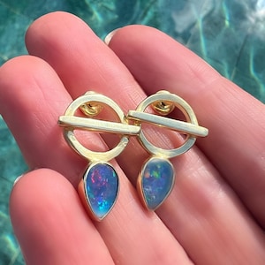 Puede incluir: Un par de pendientes dorados. Cada pendiente presenta una parte superior circular con una barra horizontal y un colgante de ópalo en forma de lágrima. Los ópalos muestran una mezcla de colores azules, morados y verdes. Los pendientes se sostienen en una mano.