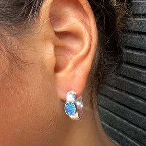 Op de afbeelding: Zilveren oorring met een ovale, blauwe opaalsteen. De oorring wordt gedragen op het oor van een persoon. De opaalsteen heeft een levendige, iriserende kwaliteit, die tinten blauw en groen weerspiegelt. De oorring is een eenvoudig, maar elegant sieraad.