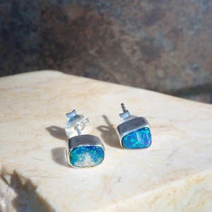 Könnte beinhalten: Ein Paar Ohrstecker mit rohen, unregelmäßig geformten Opalen mit leuchtend blauen und grünen Blitzen, gefasst in polierten Silberfassungen. Die Ohrringe verfügen über standardmäßige Silberstecker und zeigen die natürliche Textur der Steine.