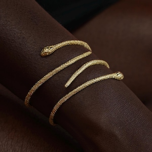 May include: A gold-toned metal bracelet shaped like a coiled snake with a textured, scale-like surface. The design features a detailed snake head at one end and a tapered tail at the other, wrapping around in a spiral pattern to form the bracelet.