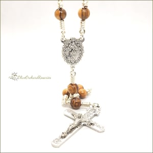 May include: A rosary with olive wood beads and silver-toned metal components. The rosary features a crucifix and a decorative centerpiece with a religious figure. The beads are a light brown color, and the metal has intricate detailing.
