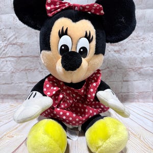 May include: A plush Minnie Mouse doll with black ears, a red polka dot bow, and a red polka dot dress. The doll has white gloves and yellow feet. The background is a light-colored brick wall and a wooden surface.