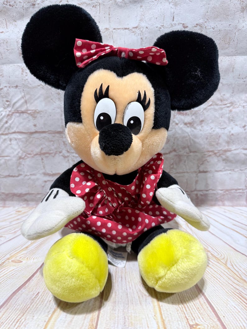 May include: A plush Minnie Mouse doll with black ears, a red polka dot bow, and a red polka dot dress. The doll has white gloves and yellow feet. The background is a light-colored brick wall and a wooden surface.