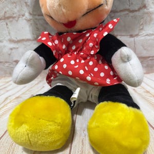 K&ouml;nnte beinhalten: Eine Minnie Mouse Pl&uuml;schpuppe. Minnie hat einen schwarzen Kopf und Ohren, ein rotes Kleid mit wei&szlig;en Punkten, wei&szlig;e Handschuhe und gelbe Schuhe. Die Puppe sitzt auf einer Holzoberfl&auml;che.