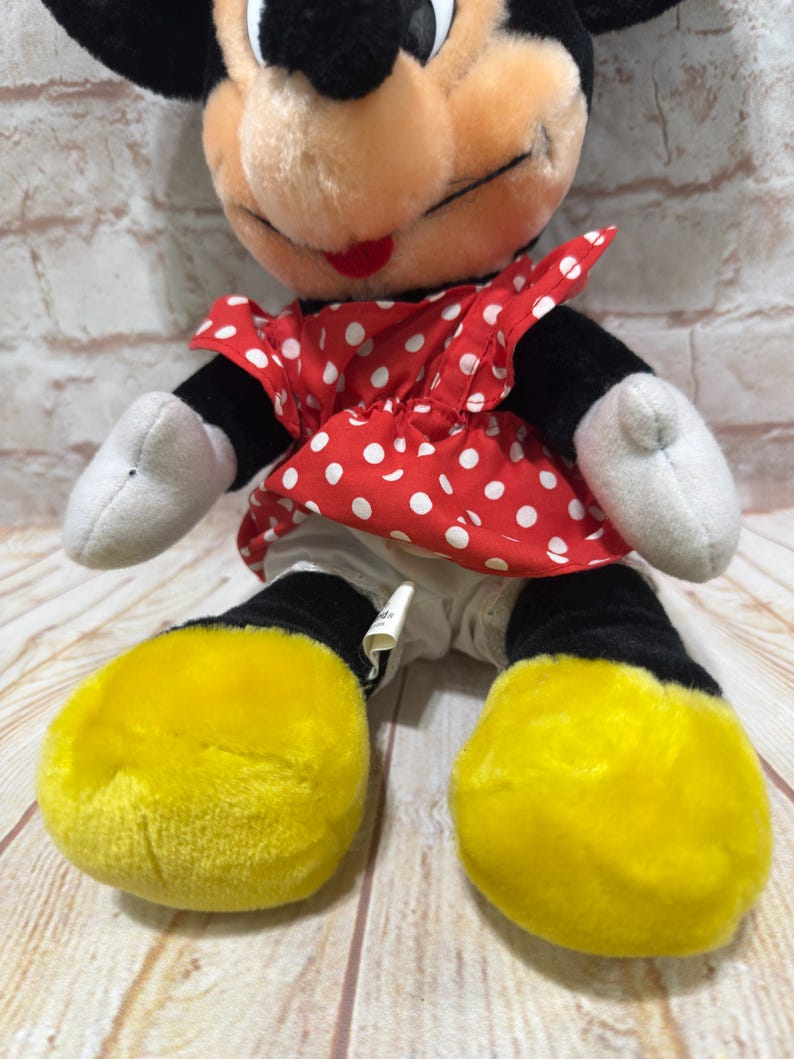 K&ouml;nnte beinhalten: Eine Minnie Mouse Pl&uuml;schpuppe. Minnie hat einen schwarzen Kopf und Ohren, ein rotes Kleid mit wei&szlig;en Punkten, wei&szlig;e Handschuhe und gelbe Schuhe. Die Puppe sitzt auf einer Holzoberfl&auml;che.