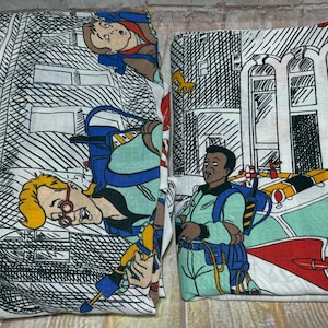 May include: Two folded fabric sheets featuring a white background with a black and white comic book style city street print. The pattern includes cartoon characters in light green jumpsuits with blue backpacks and a white vehicle with red accents.
